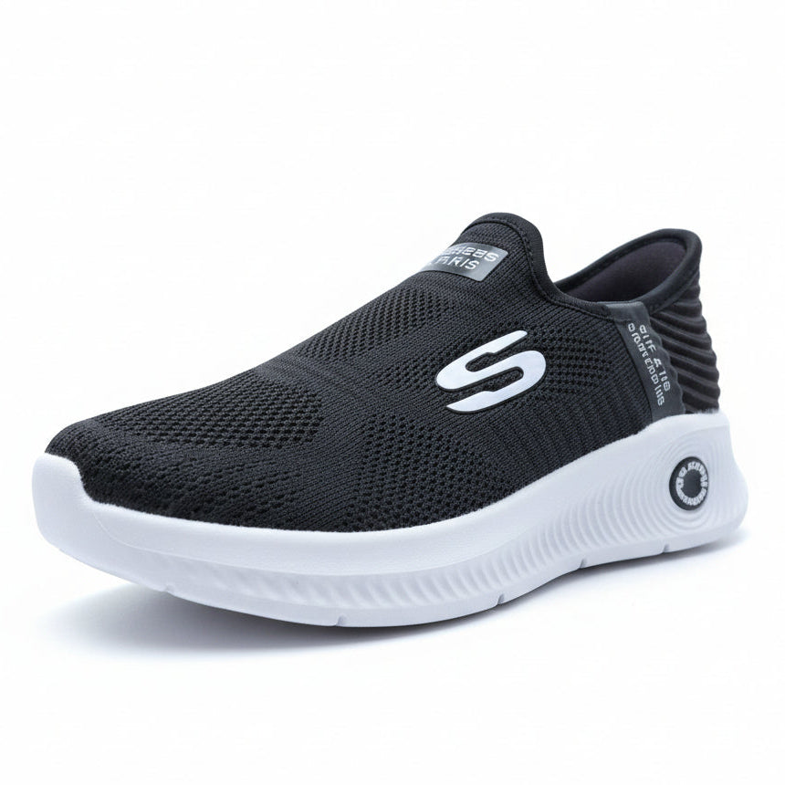 Black slip-on sneakers with white soles on a textured surface with blurred greenery in the background